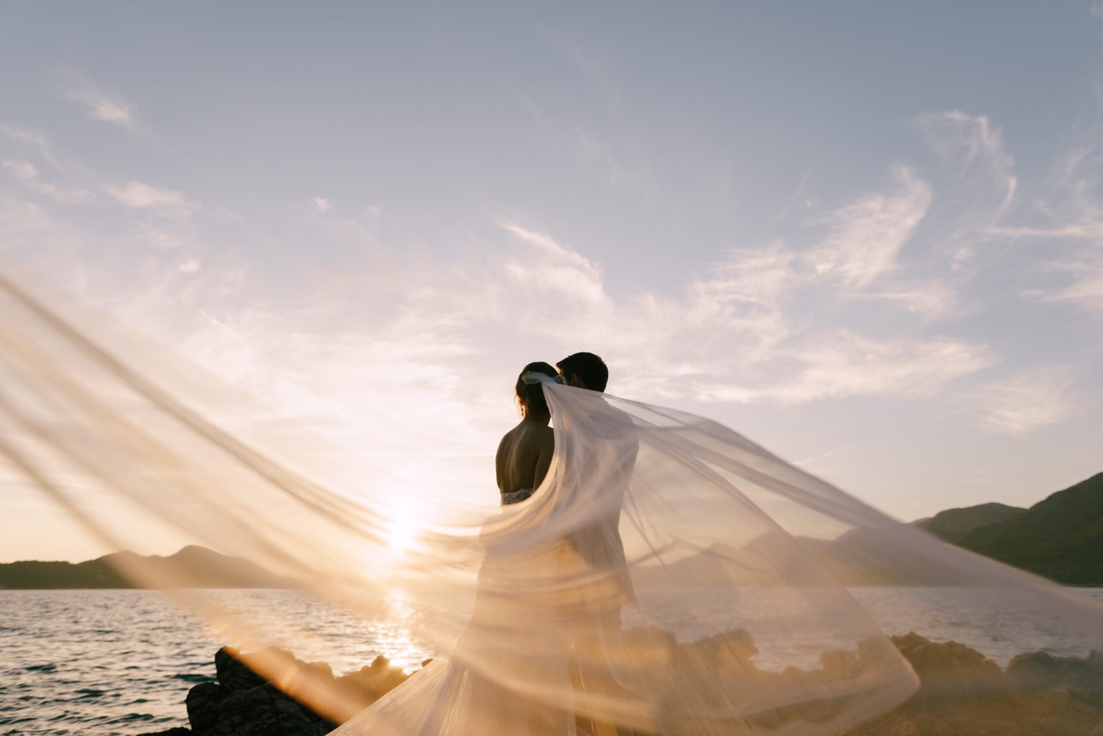 Groom hugs bride in a veil fluttering in the wind on the rocky seashore